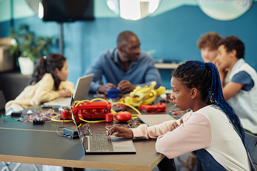 Side view portrait of young black girl using laptop in engineering class and programming robots, copy space