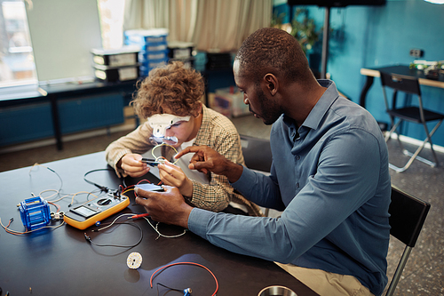 High angle portrait of young boy building robots in engineering class with male teacher helping