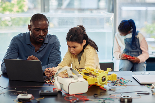 Portrait of Asian teenage girl building robots in engineering class and using laptop with male teacher helping, copy space