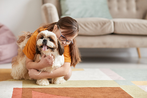 Minimal full length portrait of teenage girl with Down syndrome playing with dog on floor in cozy home interior, copy space