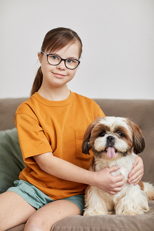 Vertical portrait of teenage girl with Down syndrome sitting with cute dog on couch and looking at camera smiling