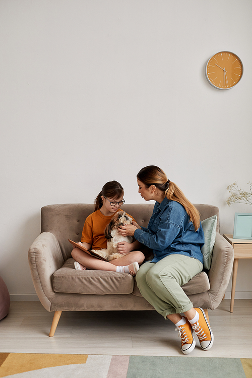 Minimal full length portrait of mother and daughter with Down syndrome playing with dog while sitting on couch at home