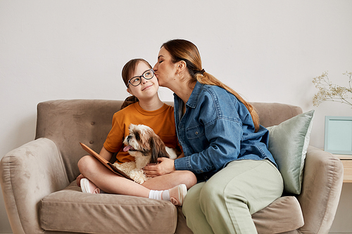 Minimal portrait of caring mother kissing daughter with Down syndrome on cheek while sitting together on couch at home