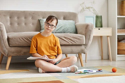 Full length portrait of teenage girl with Down syndrome sitting on floor at home cross legged and looking at camera, copy space