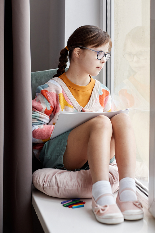 Vertical full length portrait of teenage girl with Down syndrome drawing pictures while sitting by window
