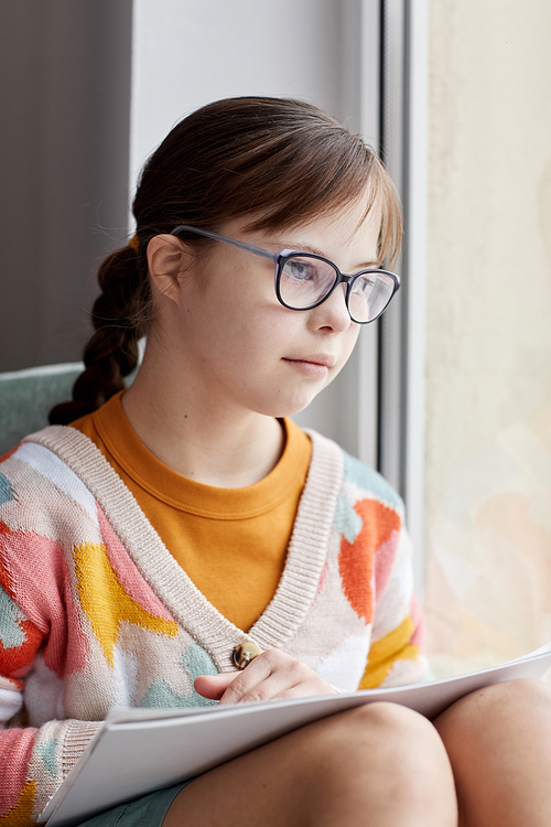 Vertical portrait of teenage girl with Down syndrome drawing pictures while sitting by window at home
