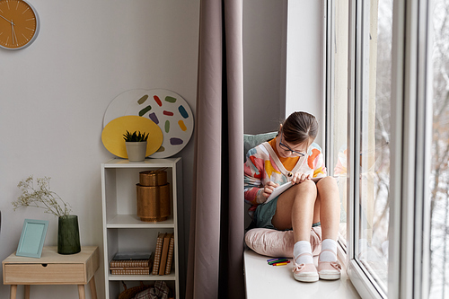Full length portrait of teenage girl drawing pictures while sitting by window in cozy room, copy space