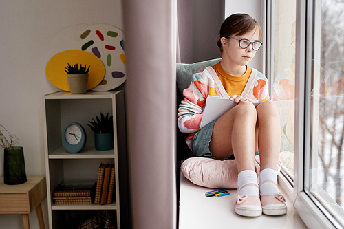 Full length portrait of teenage girl with Down syndrome drawing pictures while sitting by window in cozy room, copy space
