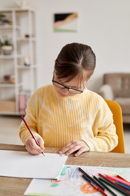 Vertical portrait of teenage girl with Down syndrome drawing pictures at table in cozy room