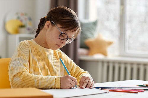 Portrait of cute girl with Down syndrome drawing pictures while sitting at desk at home, copy space