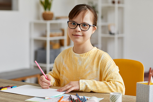Portrait of cute teen girl with Down syndrome drawing pictures while sitting at desk and smiling at camera