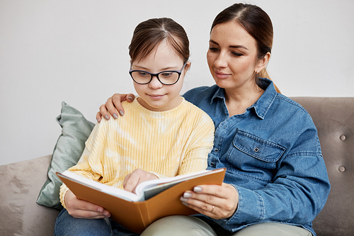 Portrait of teen girl with Down syndrome reading book with loving mother at home