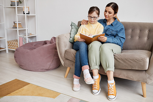 Full length portrait of teenage girl with Down syndrome reading book with loving mother at home