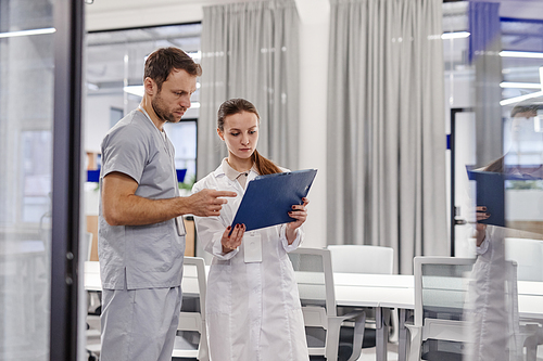 Young female general practitioner in lab coat showing medical document to colleague while consulting with him about information