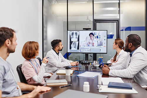 Large group of intercultural medical workers looking at their colleague on computer screen while watching online presentation
