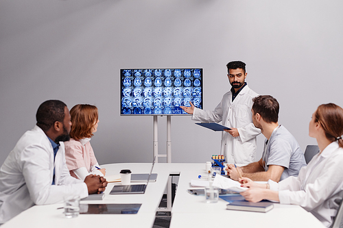 Confident oncologist in lab coat making presentation of patient mri scan to young intercultural colleagues at meeting in boardroom