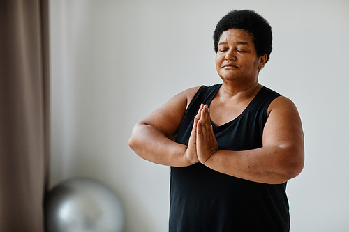Waist up portrait of black senior woman meditating with eyes closed while doing yoga indoors
