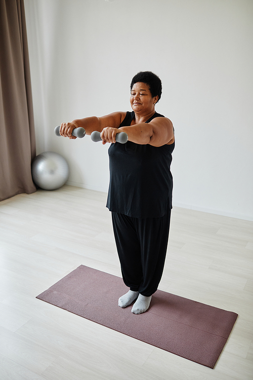 Vertical full length portrait of active senior woman lifting dumbbells while working out indoors