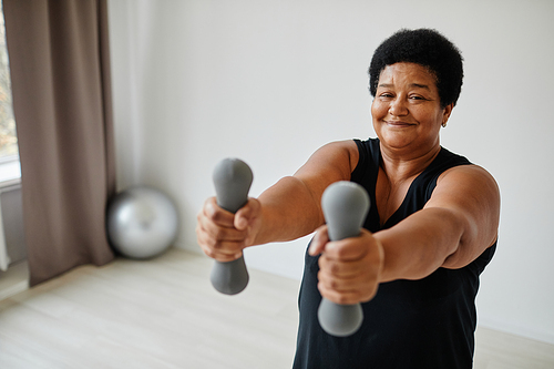 Waist up portrait of active senior woman lifting dumbbells while working out indoors and smiling at camera