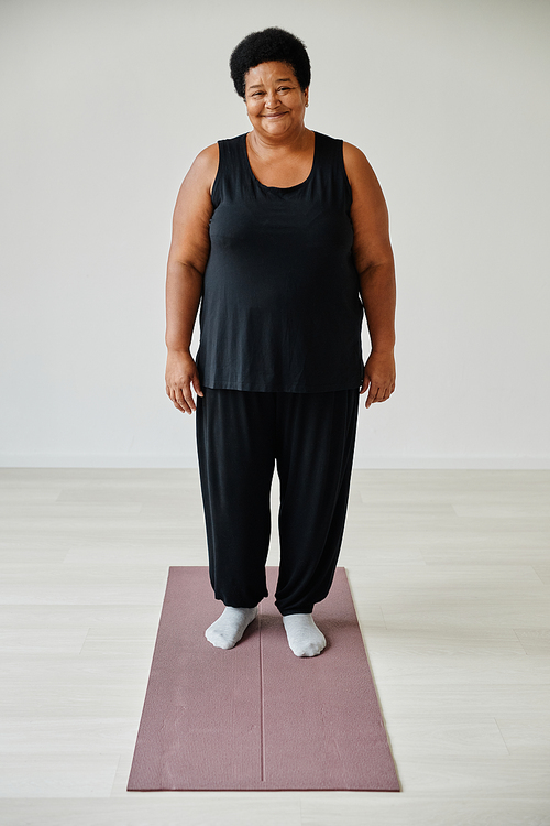 Vertical full length portrait of smiling senior woman standing on yoga mat while working out indoors