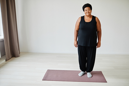 Full length portrait of smiling senior woman standing on yoga mat while working out indoors, copy space