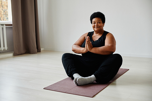Minimal full length portrait of active senior woman sitting in lotus position while enjoying yoga indoors and smiling at camera