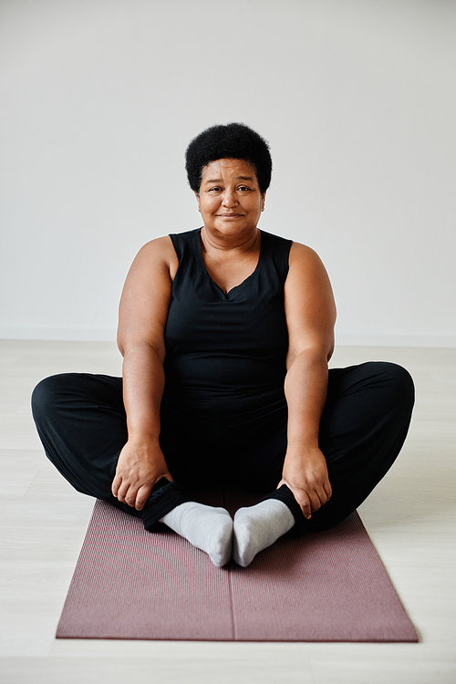 Vertical full length portrait of active senior woman sitting in lotus position while enjoying yoga indoors and smiling at camera