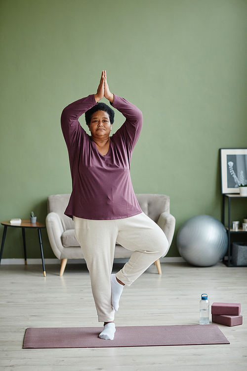 Vertical full length portrait of active senior woman doing yoga at home and practicing balance
