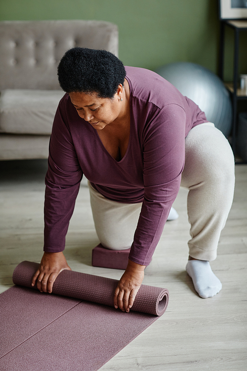 Vertical full length portrait of black senior woman unrolling yoga mat for home workout