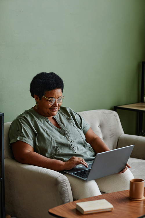 Vertical portrait of black senior woman using laptop at home while relaxing on couch