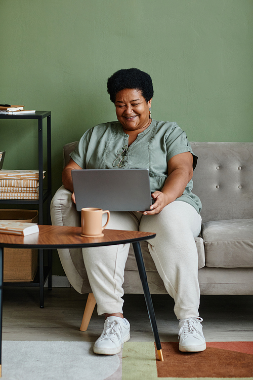 Vertical full length portrait of black senior woman using laptop at home while relaxing on couch