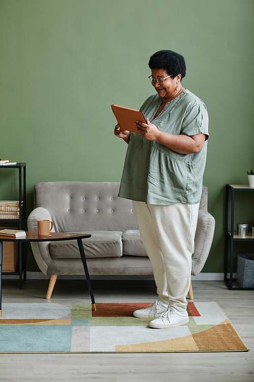 Vertical full length portrait of black senior woman using digital tablet in modern home interior