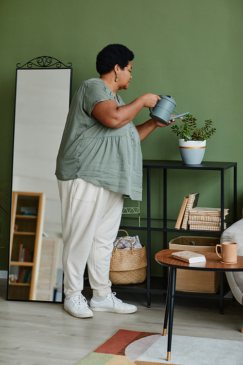 Vertical full length portrait of black senior woman watering houseplants at home