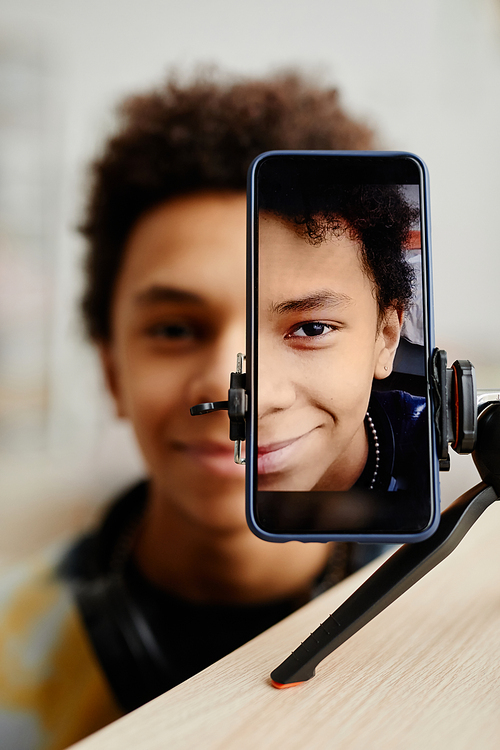 Vertical split portrait of smiling black teenager filming video for social media, focus on smartphone screen on tripod