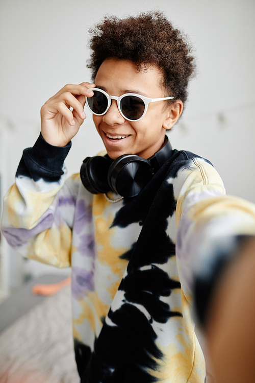 Vertical POV shot of trendy black teenager wearing sunglasses and tie dye shirt while posing for selfie photo