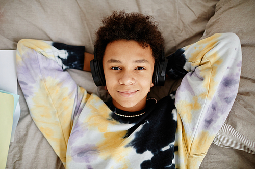 Top view portrait of black teenage boy wearing headphones and lying on bed smiling at camera