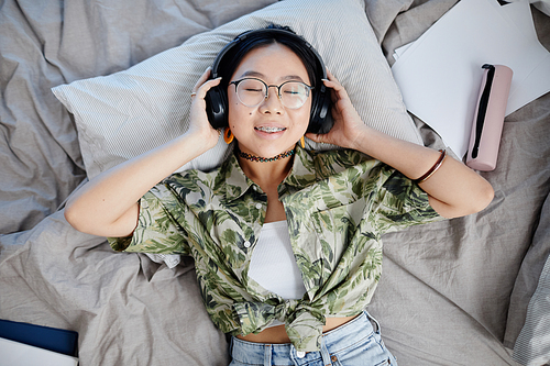Top view portrait of Asian teenage girl wearing headphones and lying on bed smiling to camera with braces