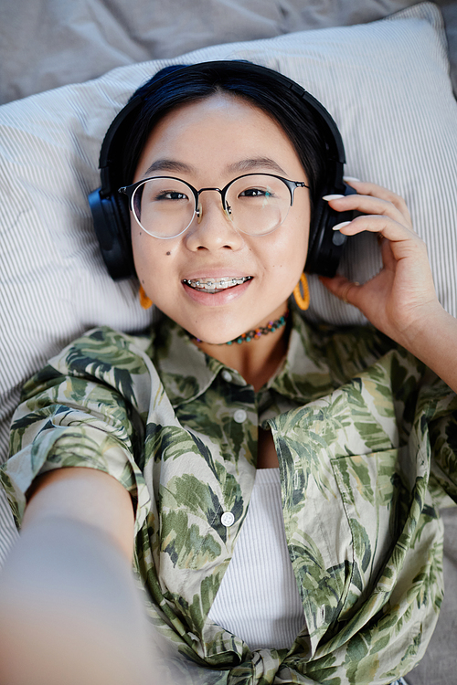Vertical POV shot of Asian teenage girl listening to music in headphones and smiling to camera while lying on bed and filming video