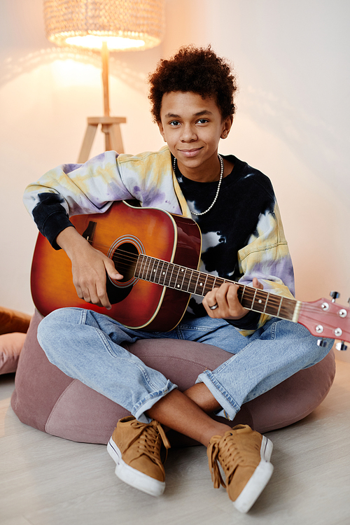 Vertical full length portrait of black teenage boy playing guitar and smiling at camera while sitting on bean bag