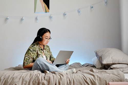 Minimal full length portrait of Asian teenage girl using laptop with headphones while sitting on bed cross legged, copy space