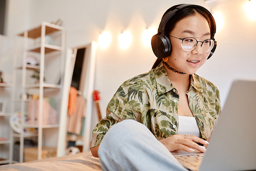 Portrait of cute Asian teenage girl wearing headphones while speaking to friend on video chat and sitting on bed, copy space