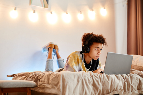 Portrait of teen African American boy using laptop while lying on bed in minimal room interior, copy space