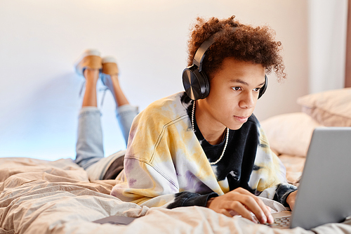 Portrait of teen black boy using computer while lying on bed in minimal room interior, copy space