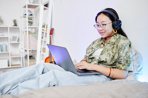 Portrait of Asian teenage girl using internet while sitting on bed in cozy room interior