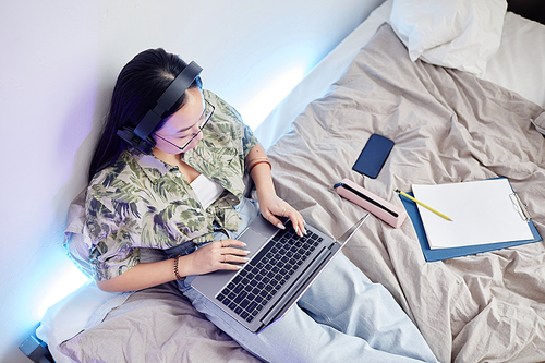 Top view portrait of Asian teenage girl using internet while sitting on bed in cozy room interior, copy space