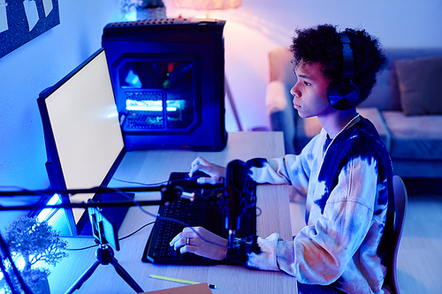 Side view portrait of black teenage boy playing video game on PC and streaming live in blue neon light