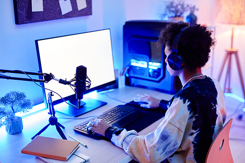 Side view of teenage boy playing video game on PC and streaming live in blue neon light with white screen mockup, copy space