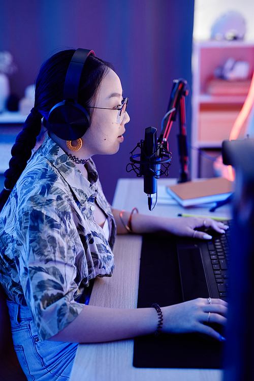 Side view of teenage girl playing video game on PC and speaking to microphone while streaming live in blue neon light