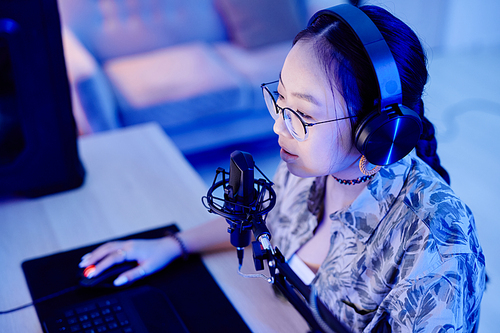 High angle portrait of young teenage girl playing video game on PC and speaking to microphone while streaming live in blue neon light