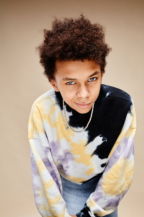 Vertical portrait of African American teenage boy wearing tie dye shirt and leaning to camera over beige background